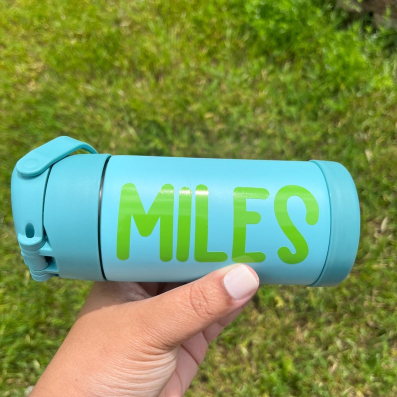 May include: A light blue water bottle with the name "MILES" in bright green letters. The bottle has a matching light blue lid and a handle. The background is green grass.