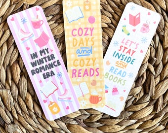 May include: Three decorative bookmarks with winter-themed designs. One bookmark has the text "IN MY WINTER ROMANCE ERA", another says "COZY DAYS and COZY READS", and the third reads "LET'S STAY INSIDE and READ BOOKS". Each bookmark features illustrations of books, drinks, and winter elements.