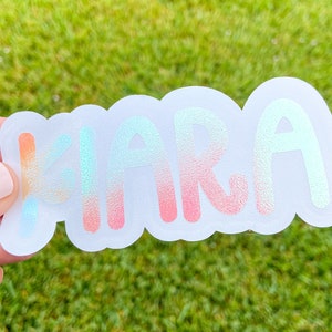 May include: A sticker with the name "Kiara" in a bubble letter font. The letters are gradient, transitioning from orange to blue and pink. The sticker has a white, cloud-shaped border.