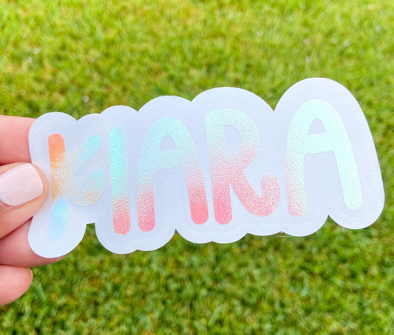May include: A sticker with the name "Kiara" in a bubble letter font. The letters are gradient, transitioning from orange to blue and pink. The sticker has a white, cloud-shaped border.