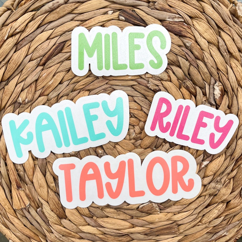 May include: Four name stickers in a rounded font with white borders. The names are "MILES" in green, "KAILEY" in turquoise, "RILEY" in pink, and "TAYLOR" in coral. The stickers are on a woven, light brown background.