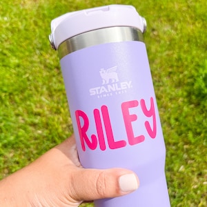 May include: A lilac Stanley tumbler with a white lid and silver details. The name "RILEY" is printed in bright pink. The Stanley logo is visible. The tumbler is designed for cold drinks.