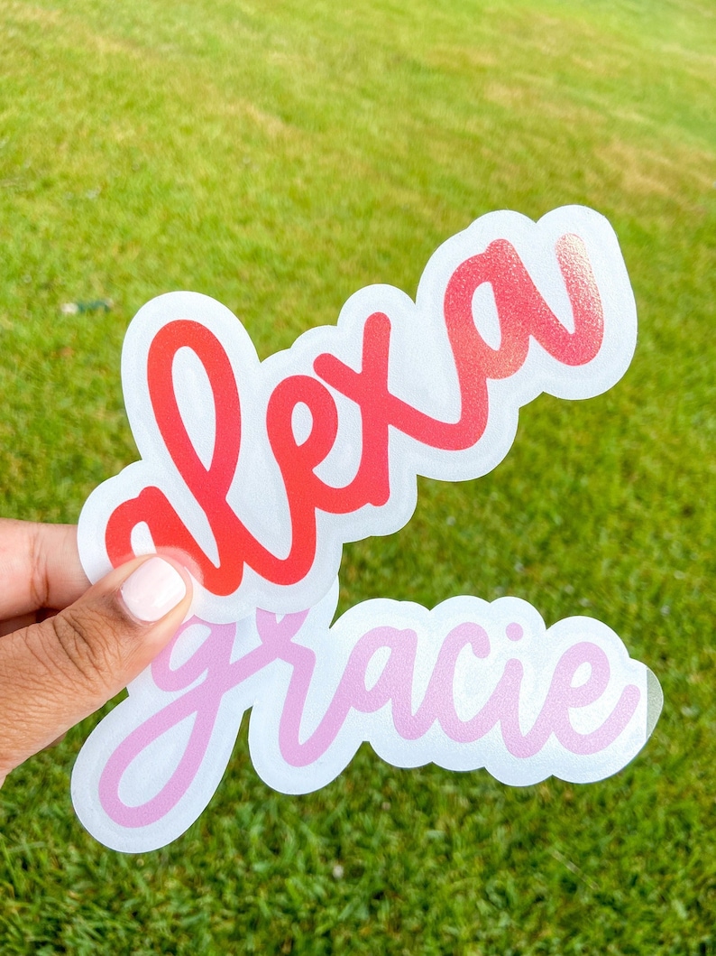 May include: Two custom name stickers, one reading "alexa" in red script and the other "gracie" in pink script. Both stickers have a white border and are held against a green grass background.