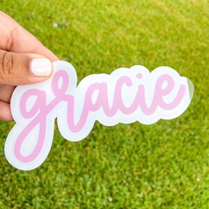 May include: A die-cut sticker with the name "Gracie" in a light pink, cursive font. The name is outlined in a darker pink and set against a white background. The sticker is held against a backdrop of green grass.