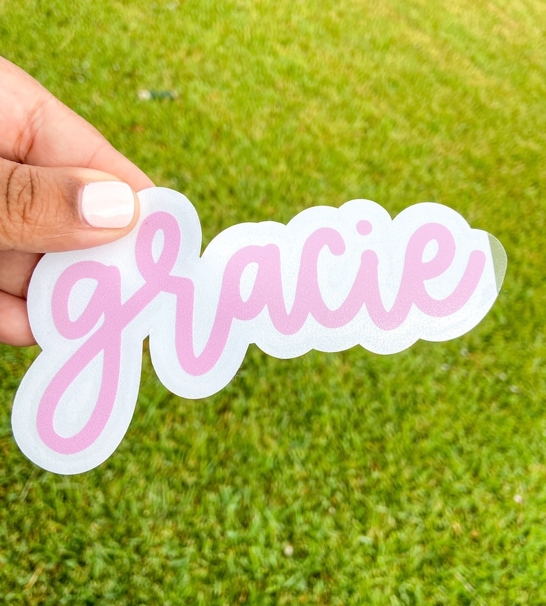 May include: A die-cut sticker with the name "Gracie" in a light pink, cursive font. The name is outlined in a darker pink and set against a white background. The sticker is held against a backdrop of green grass.