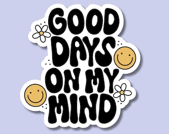 May include: A white sticker with the phrase "GOOD DAYS ON MY MIND" in bold, black, bubble letters. The design includes two yellow smiley faces and two small white flowers with yellow centers. The sticker has a light purple background.