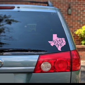 May include: A pink vinyl decal of the state of Texas with the words "Texas girl" in cursive script. The decal is affixed to the rear window of a silver vehicle. The vehicle's taillights are red.