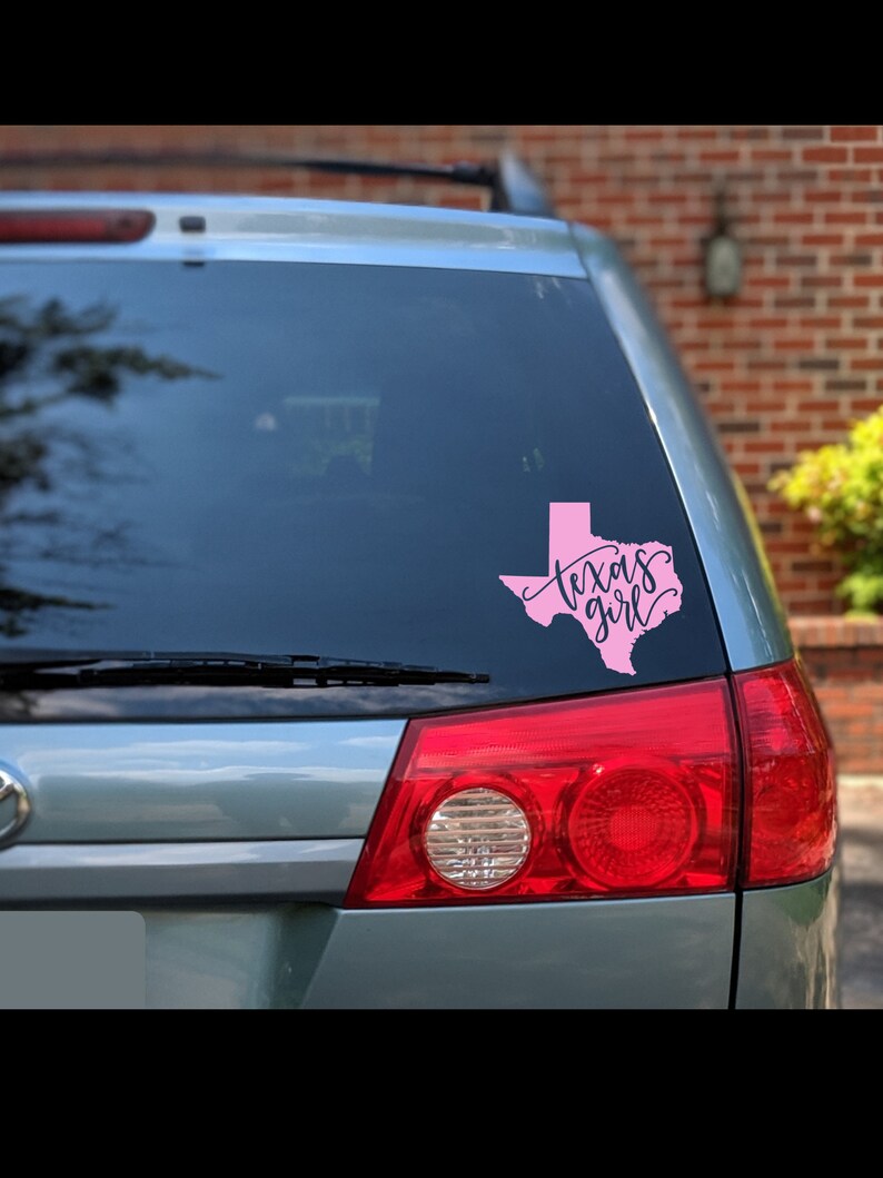 May include: A pink vinyl decal of the state of Texas with the words "Texas girl" in cursive script. The decal is affixed to the rear window of a silver vehicle. The vehicle's taillights are red.