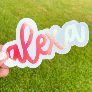 May include: A die-cut sticker with the name "alex" in a cursive font. The letters transition from red to gold with a holographic effect. The sticker is held against a green grassy background.