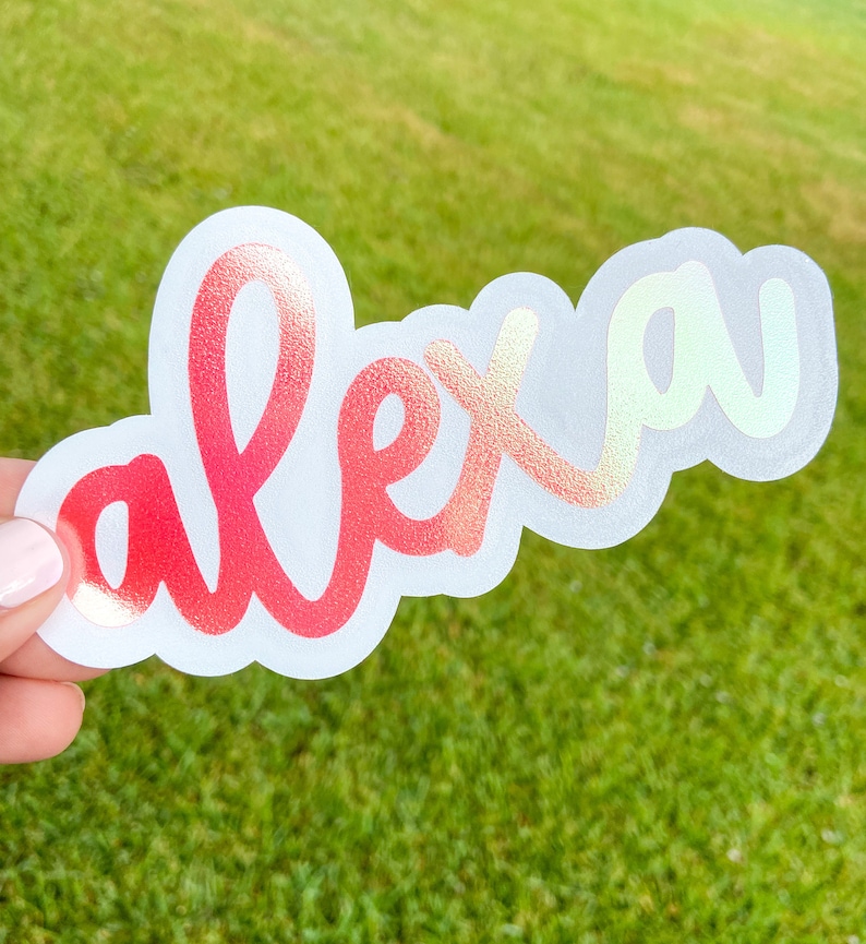 May include: A die-cut sticker with the name "alex" in a cursive font. The letters transition from red to gold with a holographic effect. The sticker is held against a green grassy background.