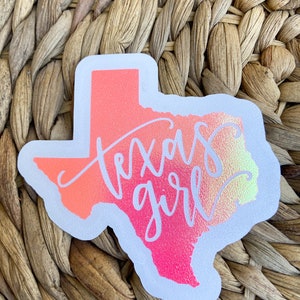 May include: A sticker in the shape of the state of Texas, with a gradient of pink, orange, and yellow. The words "Texas girl" are written in a cursive font across the state. The sticker has a white border.
