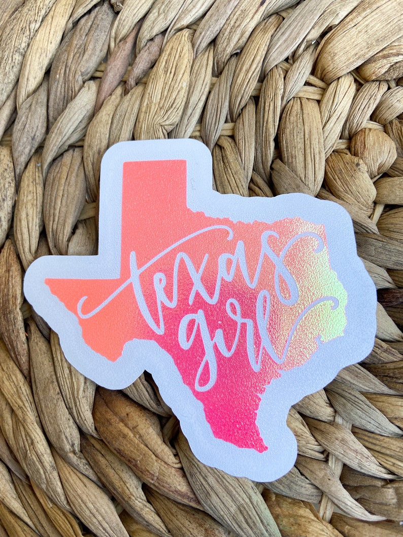May include: A sticker in the shape of the state of Texas, with a gradient of pink, orange, and yellow. The words "Texas girl" are written in a cursive font across the state. The sticker has a white border.
