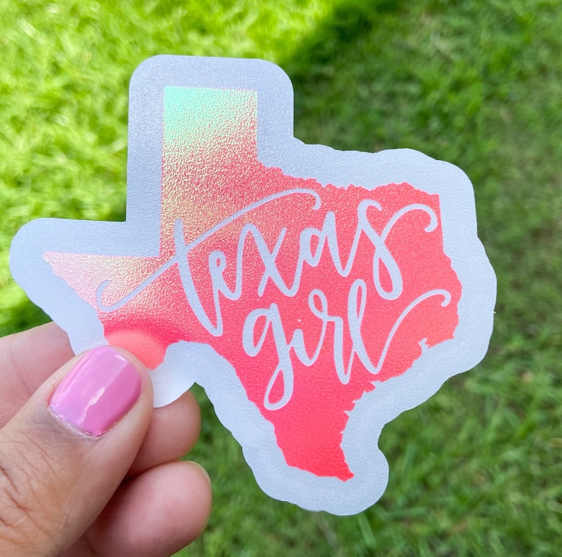 May include: A holographic sticker in the shape of Texas with the words "Texas Girl" in white script. The sticker has a gradient of pink and orange colors. The background is green grass.