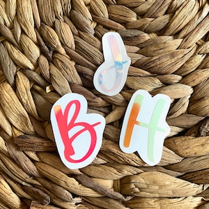 May include: Three colorful stickers with the letters "B", "J", and "H" on a woven background. The "B" is red, the "J" is a mix of colors, and the "H" is green and orange. The stickers are on a white background.