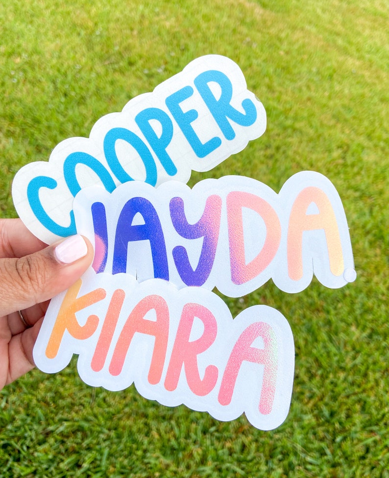 May include: Three custom name stickers with a white border. The top sticker says "COOPER" in blue. The middle sticker says "JAYDA" in purple and pink. The bottom sticker says "KIARA" in orange and pink. The stickers are held against a green grass background.