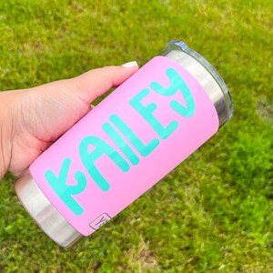 May include: A pink tumbler with the name "KAILEY" in turquoise lettering. The tumbler has a stainless steel base and a clear lid. The background is blurred green grass.