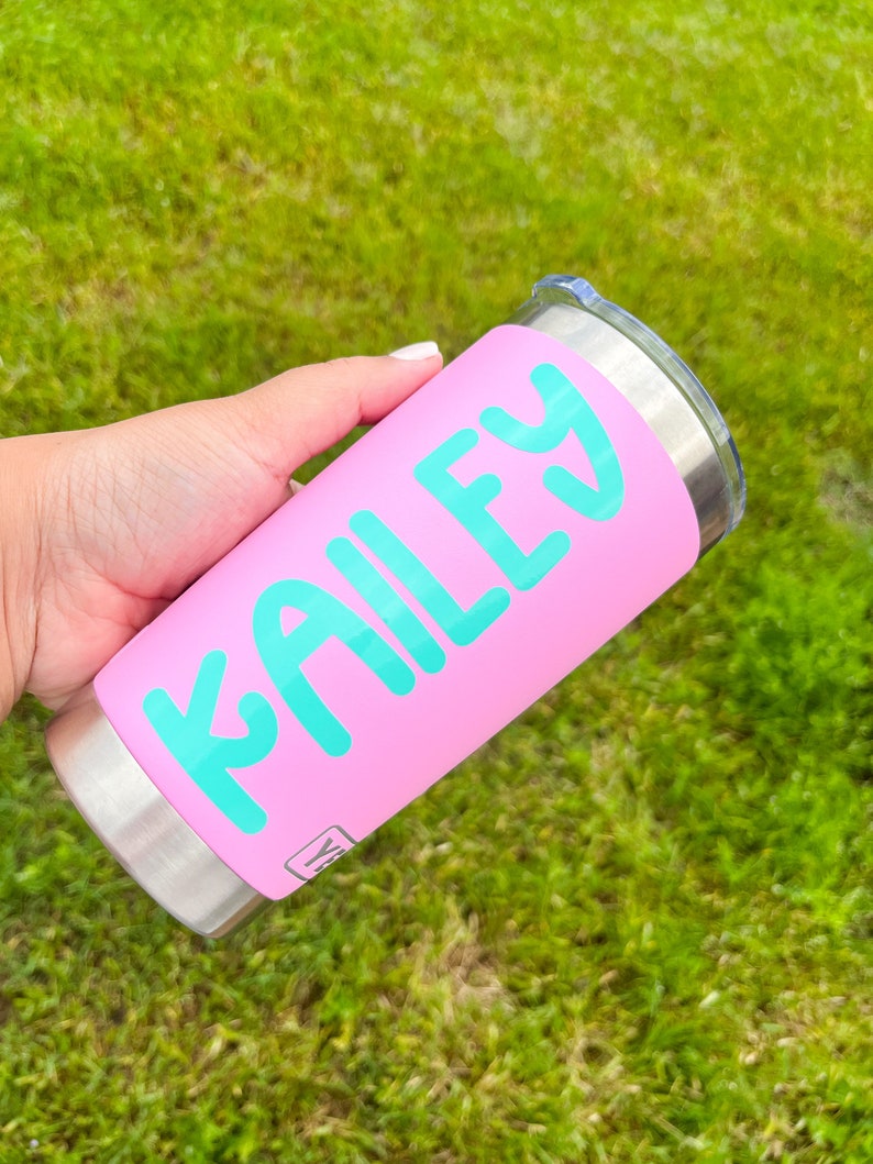 May include: A pink tumbler with the name "KAILEY" in turquoise lettering. The tumbler has a stainless steel base and a clear lid. The background is blurred green grass.