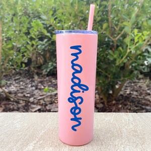 May include: A pink insulated tumbler with a blue lid and straw. The word "madison" is written vertically in blue script on the side of the tumbler. The tumbler is cylindrical and appears to be made of stainless steel.