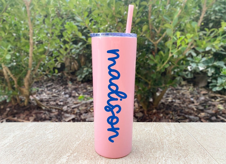May include: A pink insulated tumbler with a blue lid and straw. The word "madison" is written vertically in blue script on the side of the tumbler. The tumbler is cylindrical and appears to be made of stainless steel.