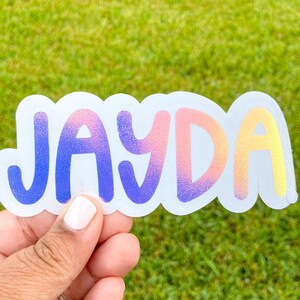 May include: A clear sticker with the name "JAYDA" in a gradient of colours, from blue to pink to yellow. The sticker has a white border and is held by a person's hand against a green background.
