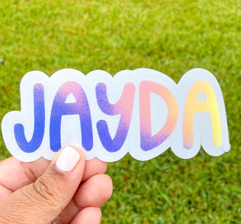 May include: A clear sticker with the name "JAYDA" in a gradient of colours, from blue to pink to yellow. The sticker has a white border and is held by a person's hand against a green background.