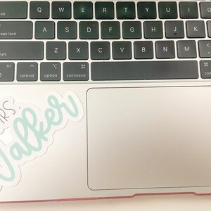 May include: A laptop keyboard with a sticker that says "MRS. Walker" in a script font. The sticker has a light blue outline and the text is in a darker blue. The keyboard is black and the laptop is silver.