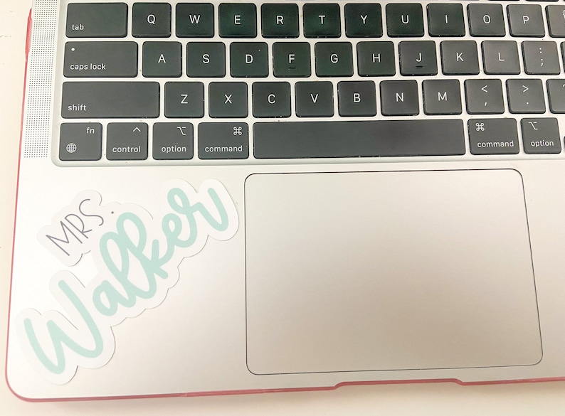 May include: A laptop keyboard with a sticker that says "MRS. Walker" in a script font. The sticker has a light blue outline and the text is in a darker blue. The keyboard is black and the laptop is silver.