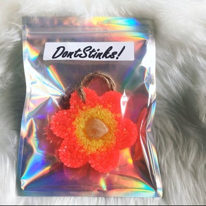 May include: A flower-shaped air freshener in a holographic bag. The air freshener is orange with yellow center beads and a clear crystal. The bag has a label that reads "DontStinks!" in black script, and a twine loop for hanging.