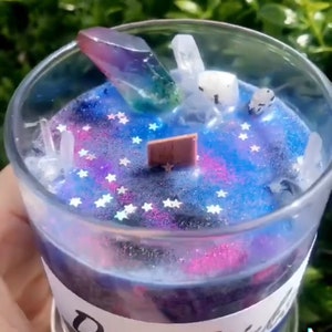 May include: A galaxy-themed candle with a clear glass jar, a purple and green crystal, and white glitter stars. The candle has a label that says "Don't Stir".