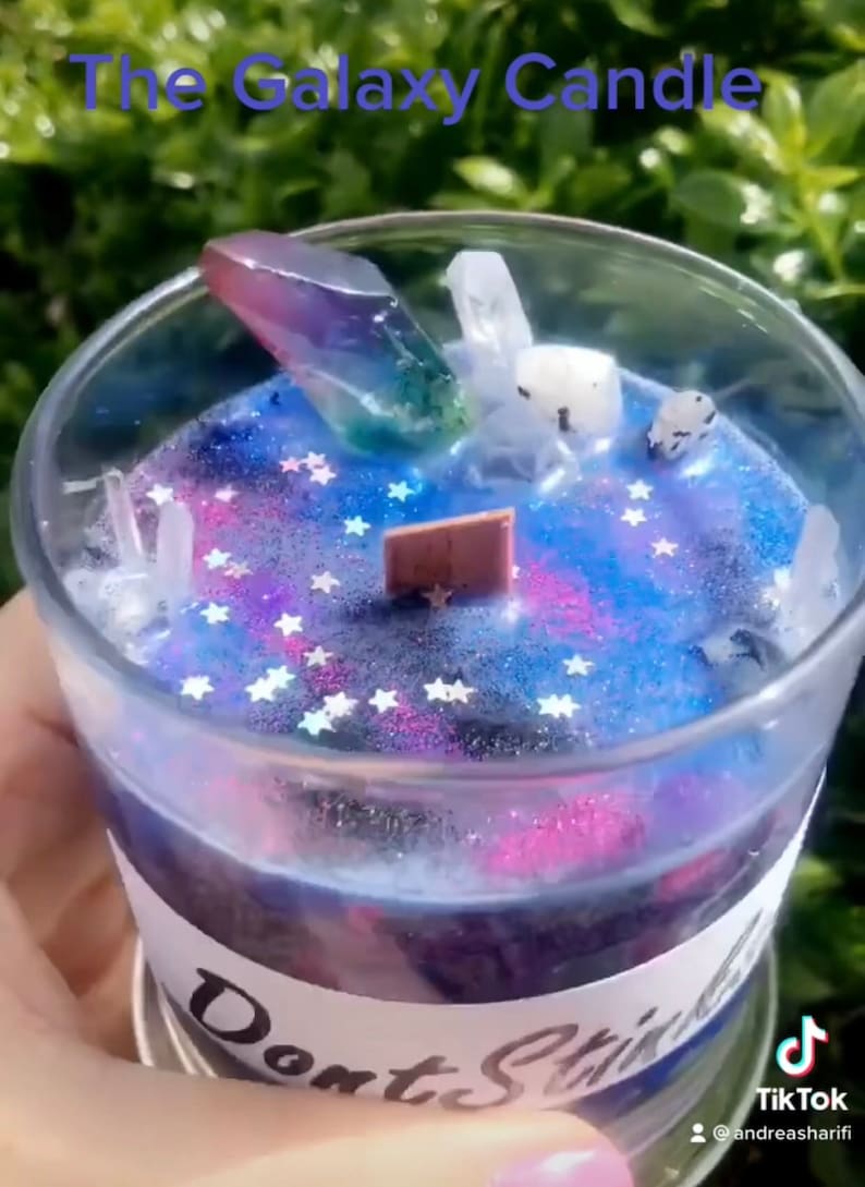 May include: A galaxy-themed candle with a clear glass jar, a purple and green crystal, and white glitter stars. The candle has a label that says "Don't Stir".