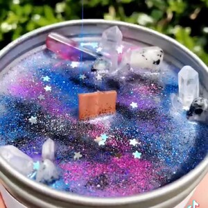 May include: A galaxy themed candle with a purple, blue, and black galaxy design. The candle is topped with clear and black crystals and silver glitter. The candle is in a silver tin with a white label that says "Don't Stop Shining".