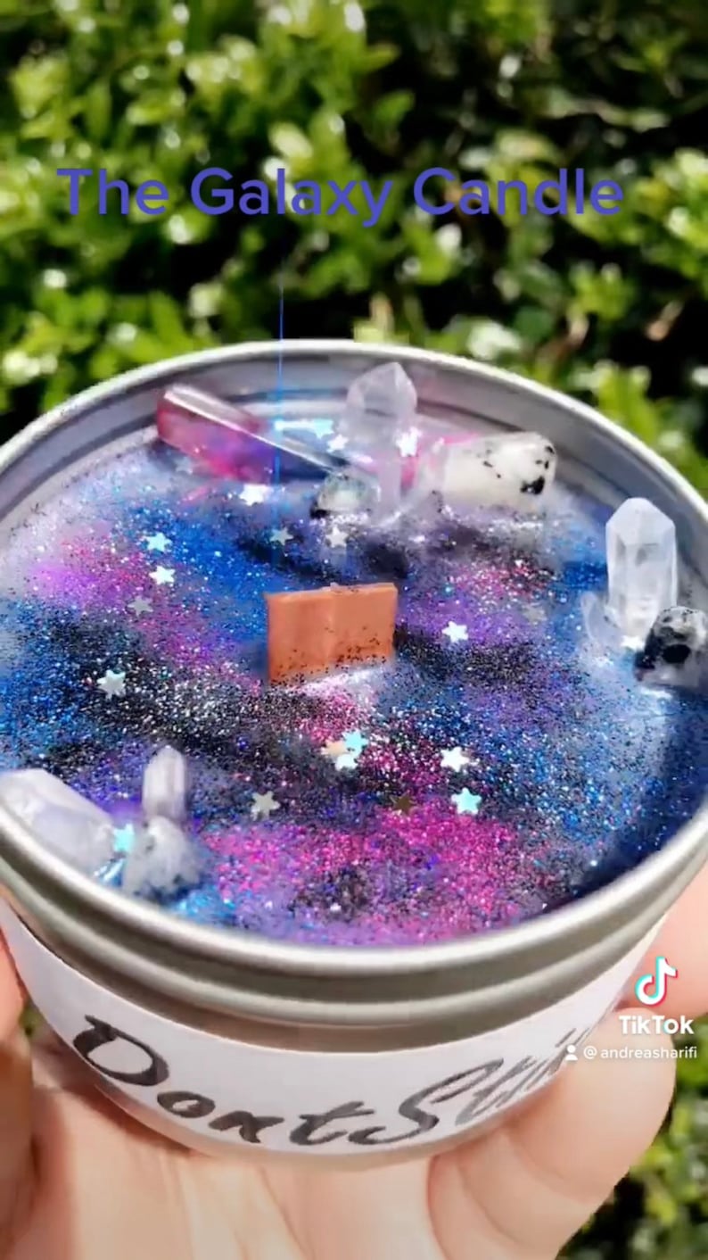 May include: A galaxy themed candle with a purple, blue, and black galaxy design. The candle is topped with clear and black crystals and silver glitter. The candle is in a silver tin with a white label that says "Don't Stop Shining".
