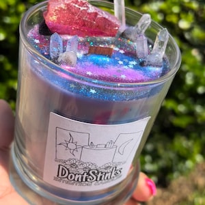 May include: A glass candle jar with a purple and blue galaxy-themed design. The jar is topped with clear crystals and a pink crystal. The label on the jar reads "DontStinks Intention Candles".