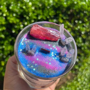 May include: A glass candle jar with a galaxy-themed design. The candle has a pink and blue glitter base with silver glitter stars. The jar contains a red crystal and several clear crystals.