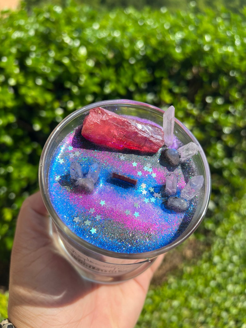 May include: A glass candle jar with a galaxy-themed design. The candle has a pink and blue glitter base with silver glitter stars. The jar contains a red crystal and several clear crystals.