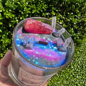 May include: A glass candle jar with a black and blue galaxy-themed design. The jar is filled with glitter and contains a pink crystal and clear quartz crystals. The label on the jar reads "Don't Shake".