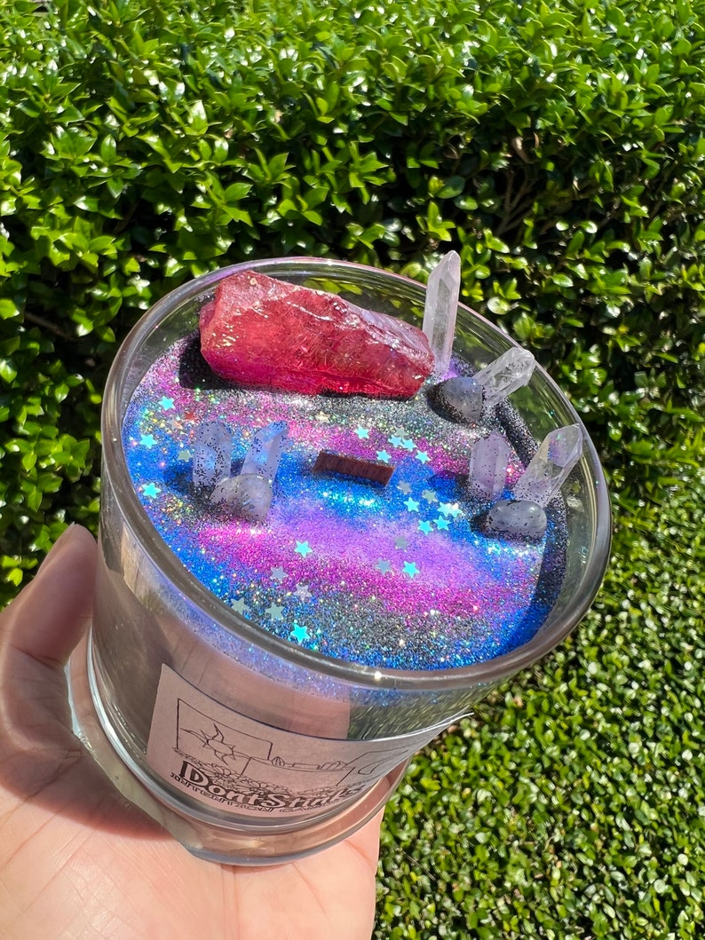 May include: A glass candle jar with a black and blue galaxy-themed design. The jar is filled with glitter and contains a pink crystal and clear quartz crystals. The label on the jar reads "Don't Shake".