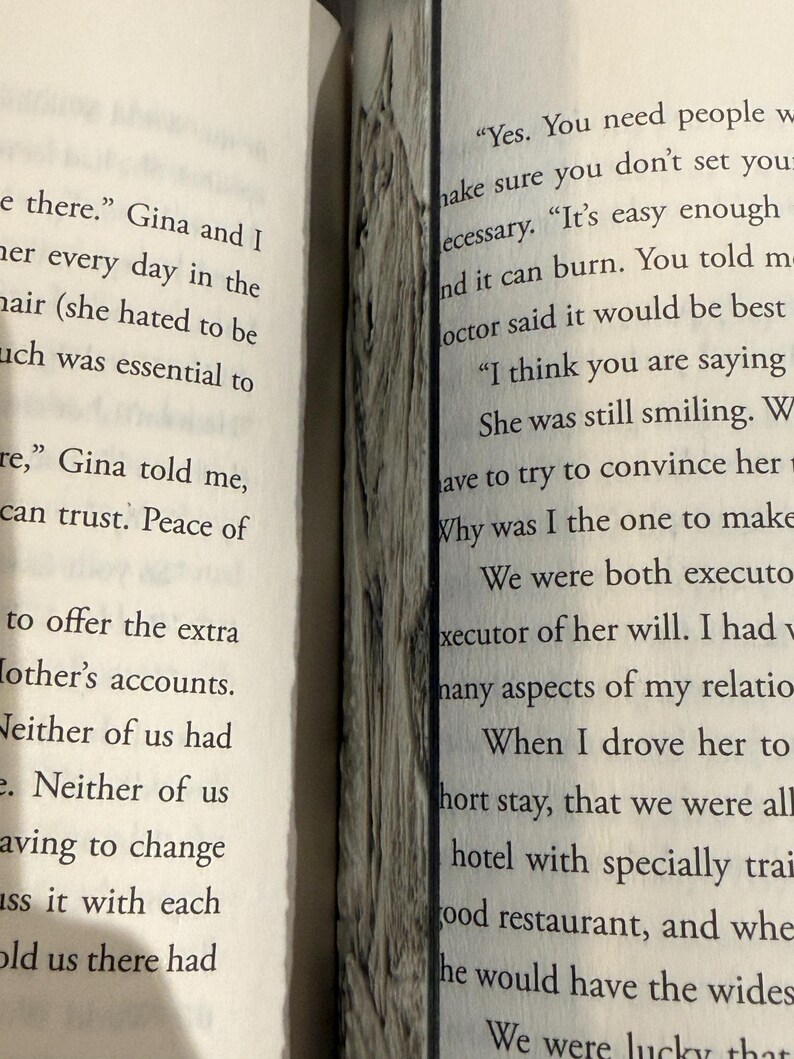 Puede incluir: Libro abierto con texto en p&aacute;ginas blancas. Un marcap&aacute;ginas decorativo de madera se coloca entre las p&aacute;ginas. El marcap&aacute;ginas tiene un aspecto texturizado gris y marr&oacute;n.
