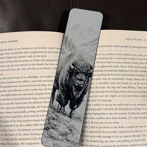 Puede incluir: Marcap&aacute;ginas rectangular con una ilustraci&oacute;n en blanco y negro de un bisonte en un paisaje natural. El marcap&aacute;ginas tiene un fondo gris claro con una cordillera en la parte superior.