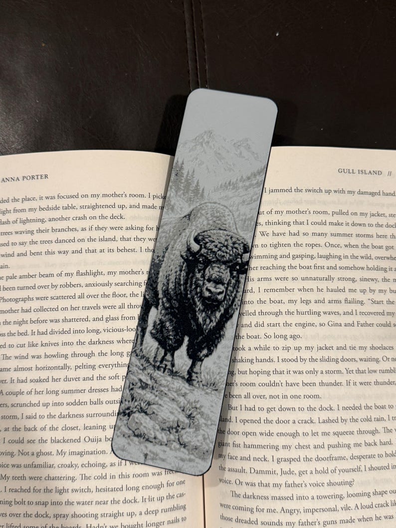 Puede incluir: Marcap&aacute;ginas rectangular con una ilustraci&oacute;n en blanco y negro de un bisonte en un paisaje natural. El marcap&aacute;ginas tiene un fondo gris claro con una cordillera en la parte superior.