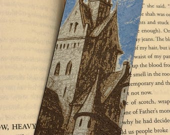 Puede incluir: Marcapáginas detallado con una ilustración de un castillo sobre un fondo de cielo azul. El castillo está representado en tonos marrones, grises y blancos, con intrincados detalles arquitectónicos. El marcapáginas está colocado en un libro abierto.
