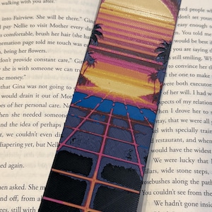 May include: A bookmark featuring a retro sunset design with a large yellow sun, palm trees, and a grid pattern. The color palette includes shades of yellow, pink, blue, and black. The bookmark is rectangular and made of a textured material.