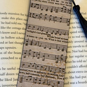 May include: A rectangular bookmark with musical notes printed on it. The bookmark has a distressed, vintage look with the text "THE STAR-" at the top. A black tassel hangs from the top. The bookmark is placed in a book.