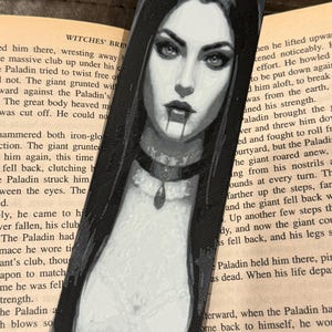 May include: Black and white bookmark featuring a gothic illustration of a woman with long dark hair, a choker, and a pendant. The woman has dark eyes and fangs, with a drop of blood. The bookmark is set against a book.
