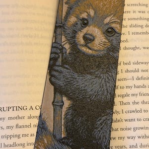 May include: A bookmark featuring a detailed illustration of a red panda holding a bamboo stalk. The red panda has reddish-brown fur with a white face and dark eyes. The bookmark is set against the backdrop of an open book.