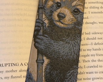 Puede incluir: Un marcapáginas con una ilustración detallada de un panda rojo sosteniendo un tallo de bambú. El panda rojo tiene pelaje marrón rojizo, una cara blanca y ojos oscuros. El marcapáginas está sobre un libro abierto.