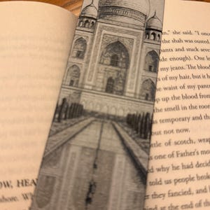 May include: A rectangular bookmark featuring a detailed black and white illustration of the Taj Mahal. The bookmark is inserted into a book, with the pages visible on either side. The image is a pencil sketch of the iconic building.