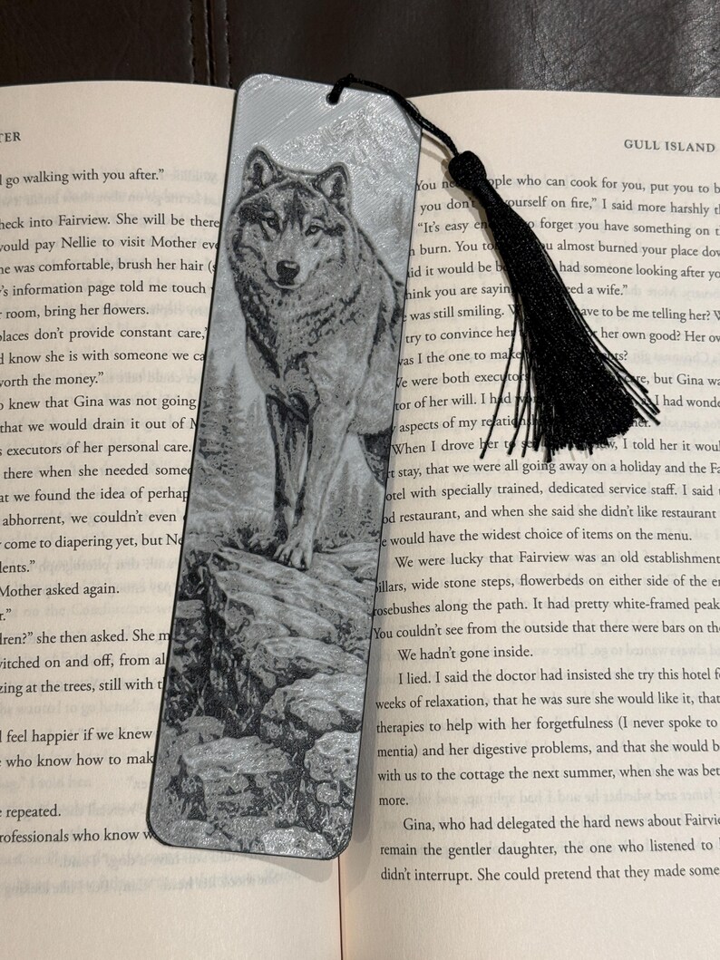 Puede incluir: Marcap&aacute;ginas plateado con un grabado detallado de un lobo sobre un terreno rocoso. El marcap&aacute;ginas tiene una borla negra y est&aacute; colocado en un libro abierto, mostrando un dise&ntilde;o cl&aacute;sico.