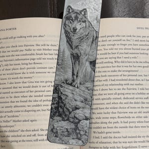 Puede incluir: Marcap&aacute;ginas de metal plateado con un grabado detallado de un lobo de pie sobre una superficie rocosa. El lobo est&aacute; mirando hacia adelante, con una representaci&oacute;n detallada de su pelaje y rasgos faciales. El marcap&aacute;ginas es rectangular.