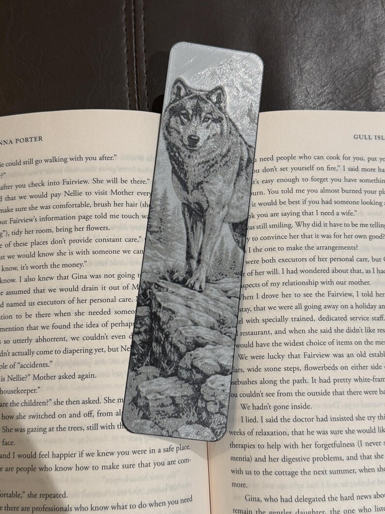 Puede incluir: Marcap&aacute;ginas de metal plateado con un grabado detallado de un lobo de pie sobre una superficie rocosa. El lobo est&aacute; mirando hacia adelante, con una representaci&oacute;n detallada de su pelaje y rasgos faciales. El marcap&aacute;ginas es rectangular.